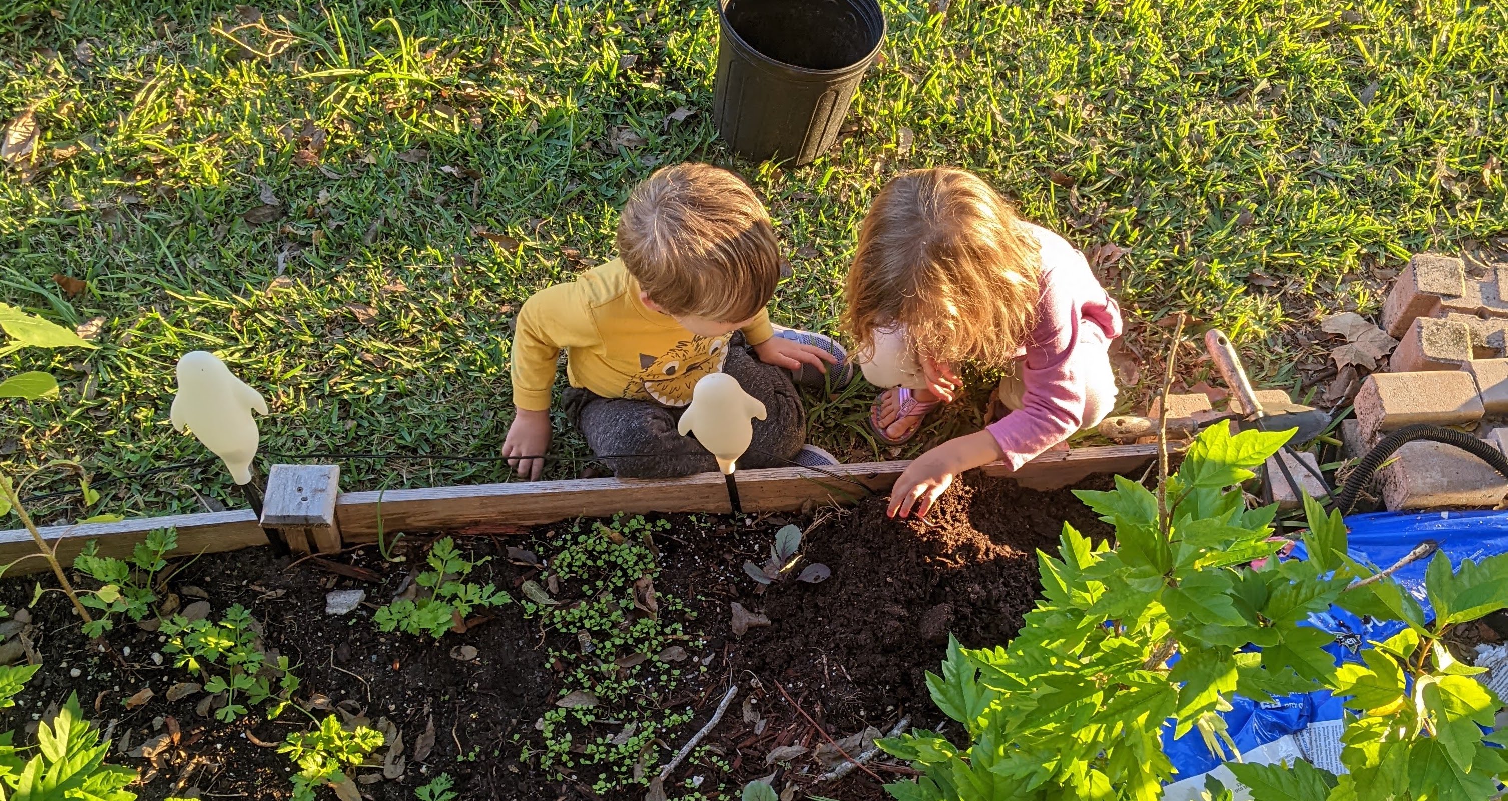 Two small children with light brown hair look down at an earthworm in dark garden soil.