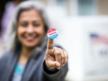 A woman with gray hair slightly blurred, holding up a finger with a voting sticker